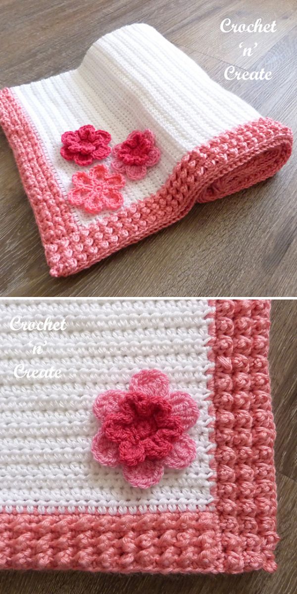 A white crocheted blanket with a pink textured border and pink flower appliqués, this charming piece is perfect for fans of crochet baby blankets, beautifully displayed on a wooden surface.