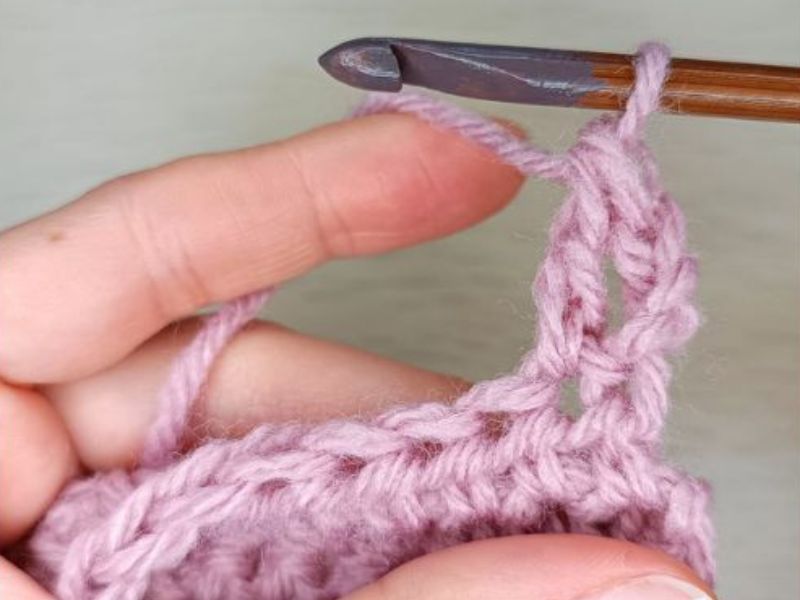 A close-up of a person's hands crocheting with pink yarn using a double crochet stitch and a wooden crochet hook.