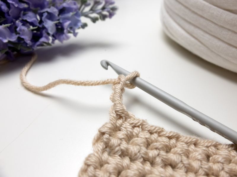 Crochet project featuring a chain stitich made with a hook and beige yarn, alongside flowers and a fabric item.