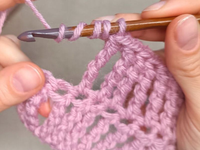 Close-up of hands demonstrating a Triple Treble Crochet Stitch tutorial with pink yarn and a wooden crochet hook, showing detailed stitches in progress.