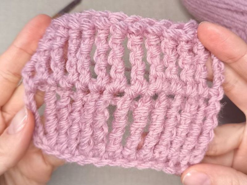 Hands holding a small swatch of pink knitted fabric, showcasing a detailed triple treble crochet stitch pattern.