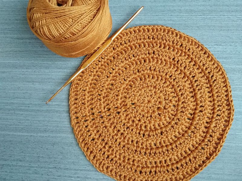 A brown crocheted placemat partially completed, with a ball of yarn and a crochet hook, on a blue wooden background.