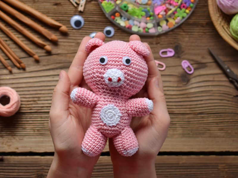 Hands holding a pink crochet teddy bear above a wooden table with crafting supplies like beads, yarn, and scissors.